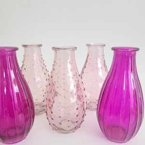 May include: A collection of glass vases in shades of pink and magenta. Two vases have vertical ribbed detailing, while others feature a textured, dotted surface. The vases are empty and have a teardrop shape.