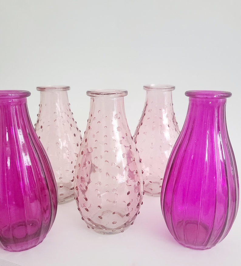 May include: A collection of glass vases in shades of pink and magenta. Two vases have vertical ribbed detailing, while others feature a textured, dotted surface. The vases are empty and have a teardrop shape.