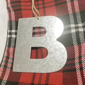 May include: A silver metal letter B ornament with a rustic finish. The ornament is hanging from a piece of twine and is on a red and black plaid background.