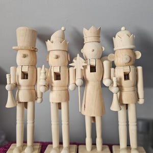 May include: Four wooden nutcrackers with different hats and accessories. One wears a top hat, another a crown, and others hold a star, a trumpet, and a small tree. Each stands on a wooden base, perfect for holiday decor.