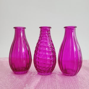 May include: Three magenta glass vases. The vases have a teardrop shape with a narrow neck. Two vases have vertical ribbed detailing, while the center vase has a textured honeycomb pattern. They are displayed on a pink surface.