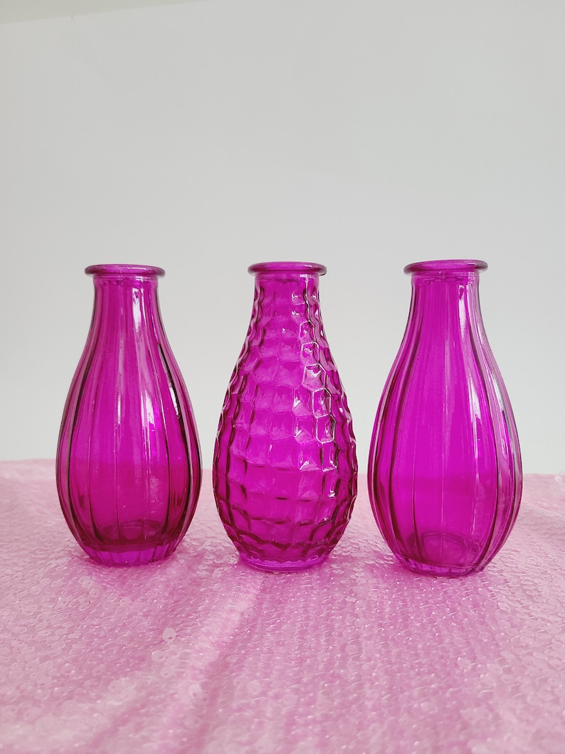 May include: Three magenta glass vases. The vases have a teardrop shape with a narrow neck. Two vases have vertical ribbed detailing, while the center vase has a textured honeycomb pattern. They are displayed on a pink surface.