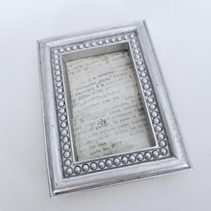 May include: A rectangular silver picture frame with a beaded border. The frame holds a piece of paper with faded text. The frame is made of a metallic material and is displayed on a white surface.