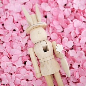 May include: A wooden toy bunny nutcracker with a white hat and a white flower in its hand. The nutcracker is standing on a bed of pink flower petals.