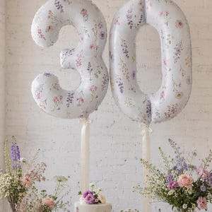 May include: Two large foil balloons shaped as the number 30, featuring a white background with a delicate multicolored wildflower print. Each balloon is attached to a cream-colored ribbon, suspended above a small cake and floral arrangements.