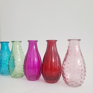 May include: A collection of five colorful glass vases in shades of turquoise, green, magenta, red, and pink. The vases have varying textures, including ribbed and dotted designs. They are arranged in a row against a white background.