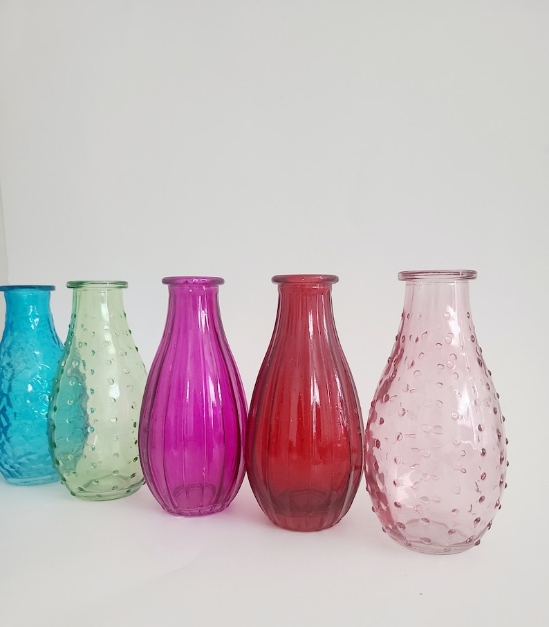 May include: A collection of five colorful glass vases in shades of turquoise, green, magenta, red, and pink. The vases have varying textures, including ribbed and dotted designs. They are arranged in a row against a white background.