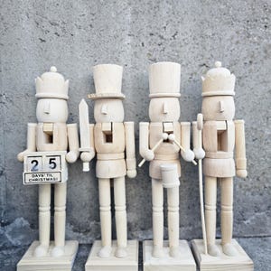 以下が含まれることがあります： 異なる楽器を持った木製のナッツクラッカーの置物が4つあります。左側のナッツクラッカーは剣を持っています。次のナッツクラッカーはドラムスティックを持っています。3番目のナッツクラッカーはドラムを持っています。最後のナッツクラッカーは杖を持っています。左側のナッツクラッカーには、25という数字と「DAYS 'TIL CHRISTMAS!」という文字が書かれた小さな木製のブロックもあります。