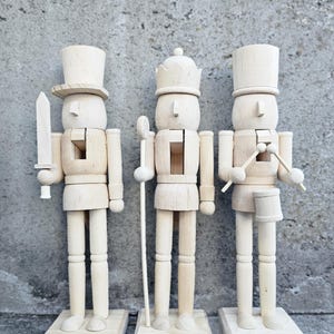 以下が含まれることがあります： 未完成の木製のナッツクラッカーの置物3つ。左のナッツクラッカーは剣を持ち、真ん中のナッツクラッカーは杖を持ち、右のナッツクラッカーはドラムスティックを持っています。