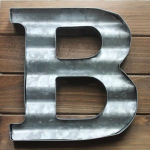 May include: A metal letter B with a corrugated surface. The letter is painted in a silver color and is mounted on a wooden background.