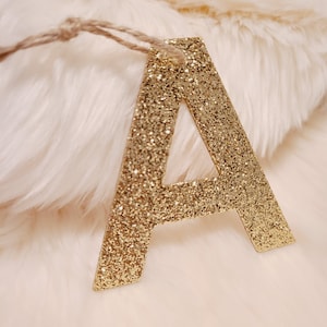 May include: Gold glitter letter A, hanging from a twine string, on a white fluffy background.