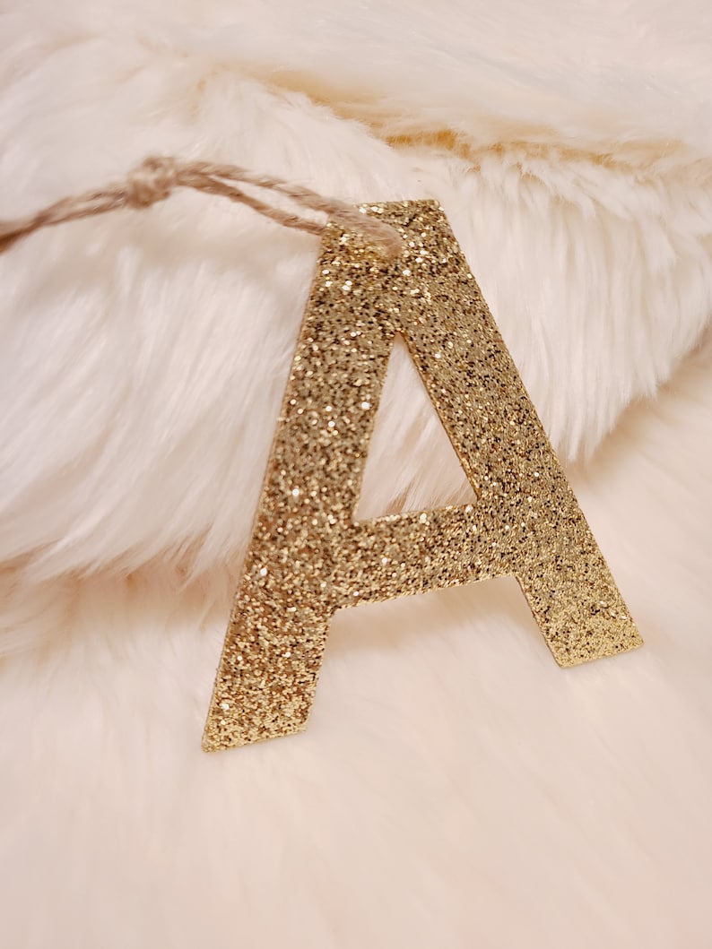 May include: Gold glitter letter A, hanging from a twine string, on a white fluffy background.
