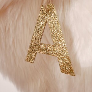 May include: A gold glitter letter A ornament, hanging from a white string. The letter is on a white fluffy background.