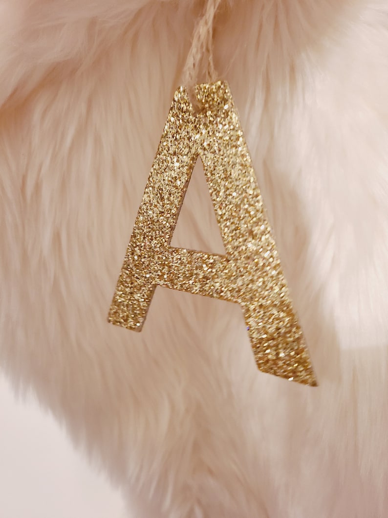 May include: A gold glitter letter A ornament, hanging from a white string. The letter is on a white fluffy background.
