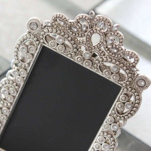 May include: A silver picture frame with a decorative, ornate design. The frame is adorned with small, clear rhinestones.