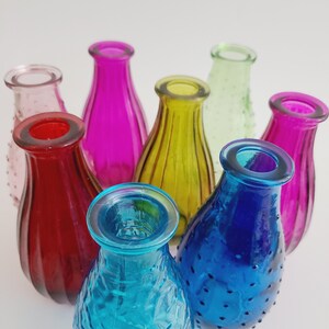 May include: A collection of small, colorful glass vases in shades of red, pink, yellow, green, blue, and purple. The vases have various textures, including smooth and textured surfaces. The vases are arranged in a circular pattern.