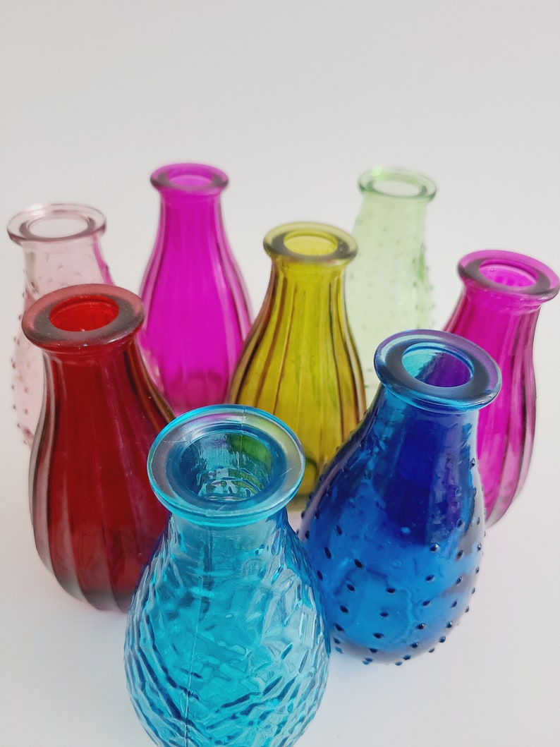 May include: A collection of small, colorful glass vases in shades of red, pink, yellow, green, blue, and purple. The vases have various textures, including smooth and textured surfaces. The vases are arranged in a circular pattern.