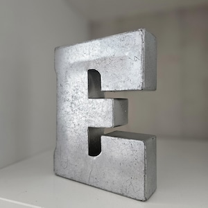 May include: A silver metal letter E with a distressed finish. The letter is standing on a white surface.