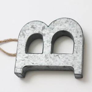 May include: A silver metal letter B with a rustic finish. The letter has a hole in the middle and is attached to a twine string.