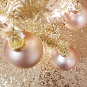 May include: Close-up of a gold Christmas tree with three rose gold ornaments. The ornaments have gold caps and are hanging from the gold branches. The background is a gold sequined fabric.