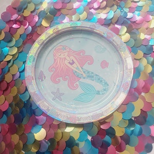 Puede incluir: Un plato de papel con temática de sirena con un fondo azul claro y una sirena con cabello rosado y cola verde. El plato tiene un borde plateado iridiscente con un diseño de concha.