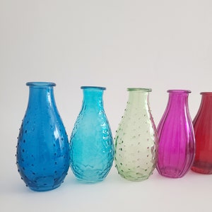 May include: A row of five colorful glass vases in shades of blue, turquoise, green, magenta, and red. The vases have a teardrop shape with textured surfaces. The background is plain white.