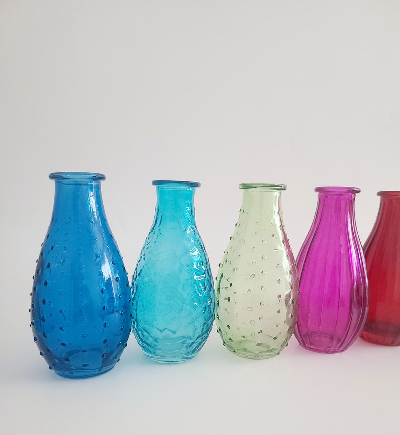 May include: A row of five colorful glass vases in shades of blue, turquoise, green, magenta, and red. The vases have a teardrop shape with textured surfaces. The background is plain white.