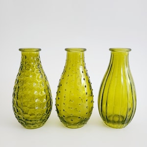 May include: Three olive green glass vases with distinct textures. The vase on the left has a honeycomb pattern, the middle one has small raised dots, and the right one has vertical ridges. Each vase has a rounded body and a narrow neck.