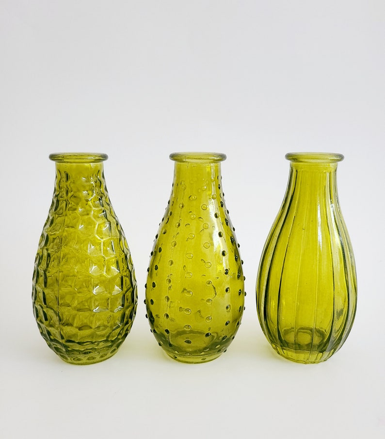 May include: Three olive green glass vases with distinct textures. The vase on the left has a honeycomb pattern, the middle one has small raised dots, and the right one has vertical ridges. Each vase has a rounded body and a narrow neck.