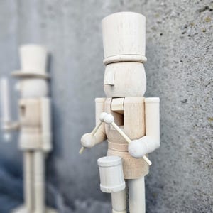 以下が含まれることがあります： 塗装されていない木製のナッツクラッカーが、ドラムスティックとドラムを持っています。ナッツクラッカーは背の高い帽子をかぶっており、顔はペイントされています。