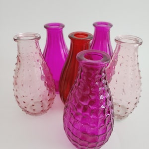 May include: A collection of six colorful glass vases in shades of pink and red. The vases have textured surfaces and a teardrop shape. The vases are arranged closely together, with a white background.