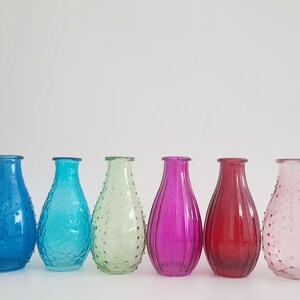 May include: Six colorful glass vases in a row. The vases are in shades of blue, turquoise, green, purple, red, and pink. Each vase has a unique texture, with some featuring a dotted pattern and others with vertical lines. The vases are empty.