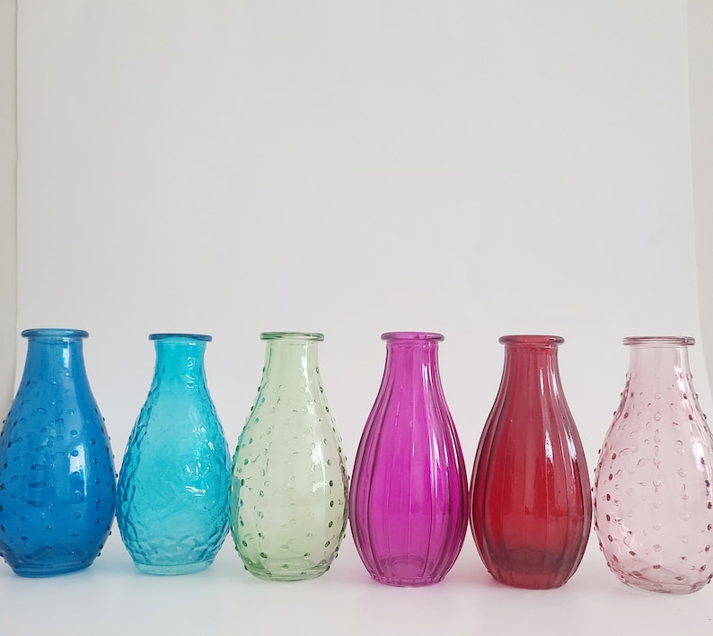 May include: Six colorful glass vases in a row. The vases are in shades of blue, turquoise, green, purple, red, and pink. Each vase has a unique texture, with some featuring a dotted pattern and others with vertical lines. The vases are empty.