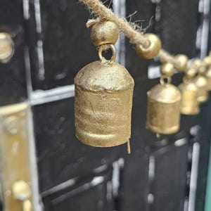 May include: A garland featuring a series of metallic gold-colored bells strung along a natural jute twine cord. Each bell is accompanied by a small gold-colored bead, creating a repeating pattern of bells and beads along the length of the rope.