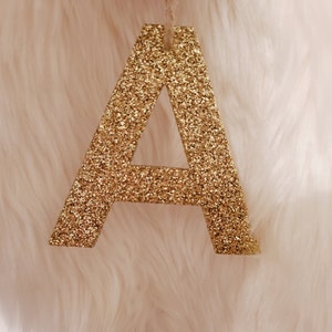 May include: A gold glitter letter A hanging from a string. The letter is against a white fuzzy background.