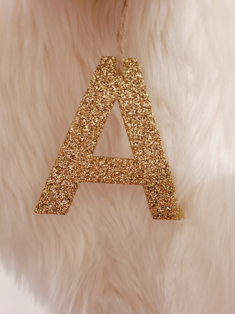 May include: A gold glitter letter A hanging from a string. The letter is against a white fuzzy background.