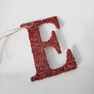 May include: A red glitter-covered letter "E" with a natural twine string attached. The letter is on a white background. The glitter gives the letter a sparkling appearance, suitable for crafts or decorations.