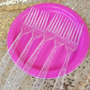 May include: Six clear plastic forks with silver glitter embedded in the plastic. The forks are arranged on a pink plate.
