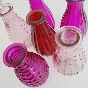 May include: A collection of small, colorful glass vases in shades of pink and red. The vases have various textures, including smooth, ribbed, and dimpled surfaces. The open tops and rounded bodies suggest they are designed for holding flowers or other decorative items.