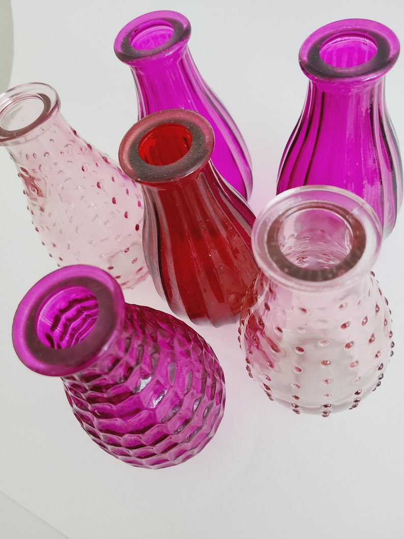 May include: A collection of small, colorful glass vases in shades of pink and red. The vases have various textures, including smooth, ribbed, and dimpled surfaces. The open tops and rounded bodies suggest they are designed for holding flowers or other decorative items.