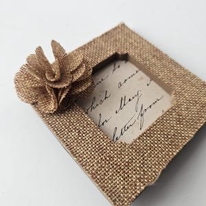 May include: A small, diamond-shaped burlap picture frame with a decorative flower on the upper left corner. The frame has a rectangular opening and contains a piece of paper with handwritten text. The frame is a natural tan color.