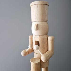 May include: Unpainted wooden nutcracker doll holding drumsticks. The nutcracker is wearing a tall hat and has a round face.