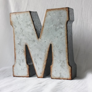 May include: A large, silver metal letter M with a rustic, galvanized finish. The letter has a slightly distressed look with some brown rust spots.