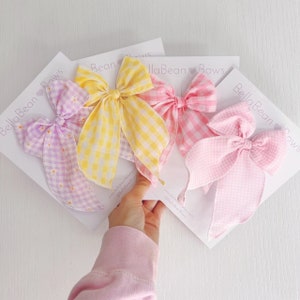 Op de afbeelding: Drie gingham haarstrikken in geel, roze en paars. De strikken zijn gebonden in een grote, pluche knoop en hebben een geruit patroon. De strikken worden gepresenteerd op witte kaarten met de tekst "BellaBean Bows" erop gedrukt.