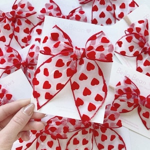 May include: A white card with red hearts printed on it. The card has the text "Bella Bean" printed on it. A red and white bow with red hearts printed on it is on top of the card.