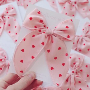 May include: A pink ribbon bow with a red heart pattern. The bow is tied and ready to be used for gift wrapping or hair accessories.