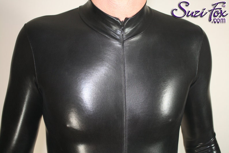 Op de afbeelding: Een zwarte, nauwsluitende bodysuit met een hoge kraag en een ritssluiting aan de voorkant. Het glanzende materiaal reflecteert licht, waardoor highlights en schaduwen ontstaan. Het Suzi Fox .com logo is zichtbaar.