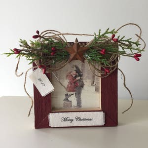 May include: A rustic red wooden picture frame with a photograph of a couple. The frame is decorated with greenery, red berries, a star, and twine. The text "Merry Christmas" is printed on a white ribbon.