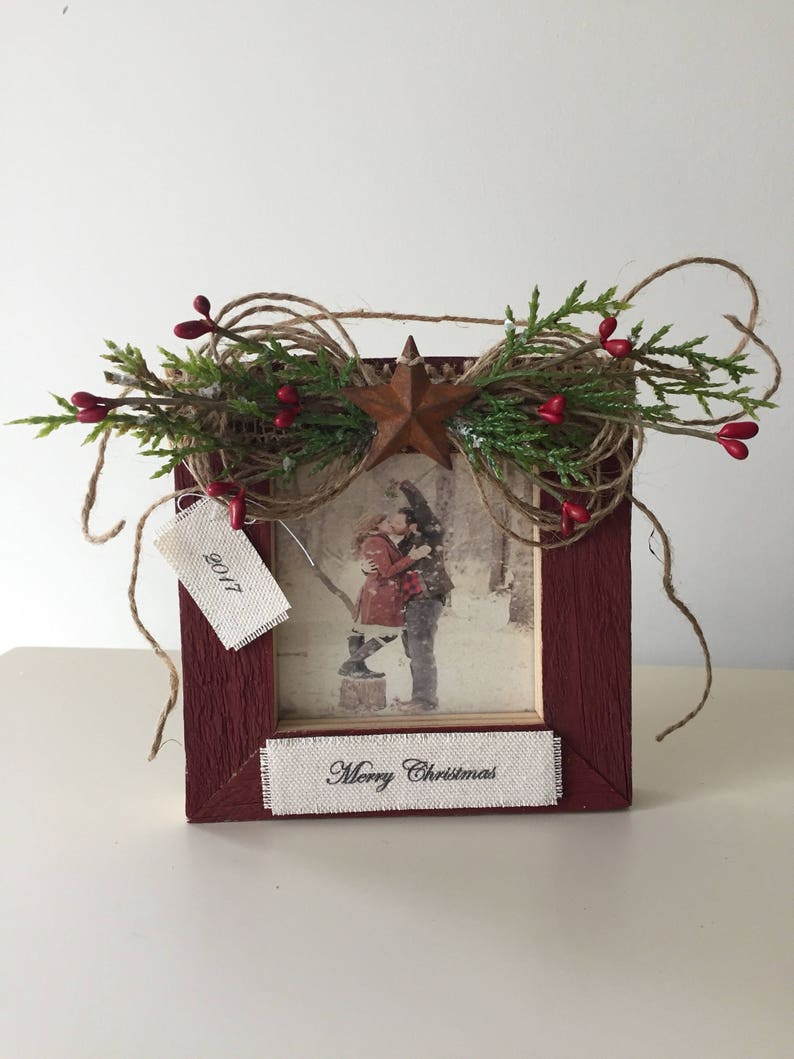 May include: A rustic red wooden picture frame with a photograph of a couple. The frame is decorated with greenery, red berries, a star, and twine. The text "Merry Christmas" is printed on a white ribbon.