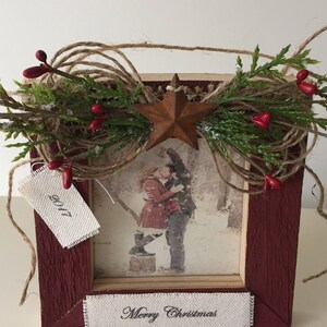 May include: A rustic red wooden picture frame decorated with greenery, red berries, and a rusty star. The frame holds a black and white photo of a couple kissing. The text "Merry Christmas" is at the bottom, and a tag reads "2017".